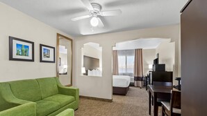 A hotel room with a green sofa, a bed, a desk with a chair, a television, and a ceiling fan.