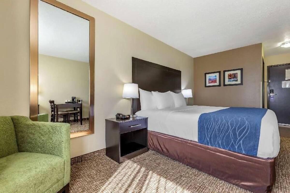 Una habitación de hotel con cama, mesita de noche, lámpara, sillón verde y un espejo grande.