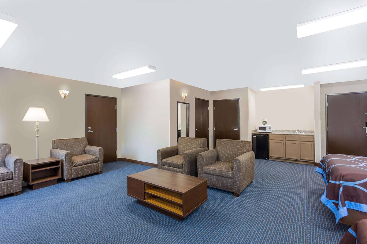 Une chambre d’hôtel avec une moquette bleue, deux fauteuils, un canapé, une table basse, une lampe, un four à micro-ondes et un lit recouvert d’une courtepointe marron et bleue.