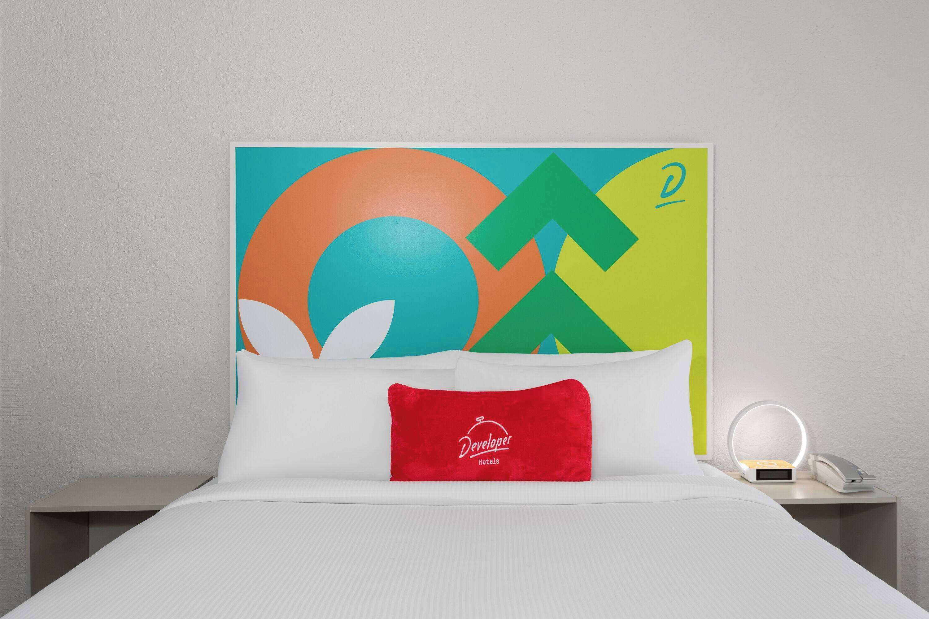 A hotel room with a bed, a red pillow with "Developer Hotels" text, a nightstand with a phone and a lamp, and a colorful abstract wall art.