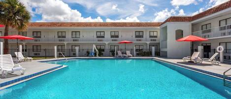 A swimming pool with lounge chairs and umbrellas, surrounded by a building with a tiled roof and balconies.
