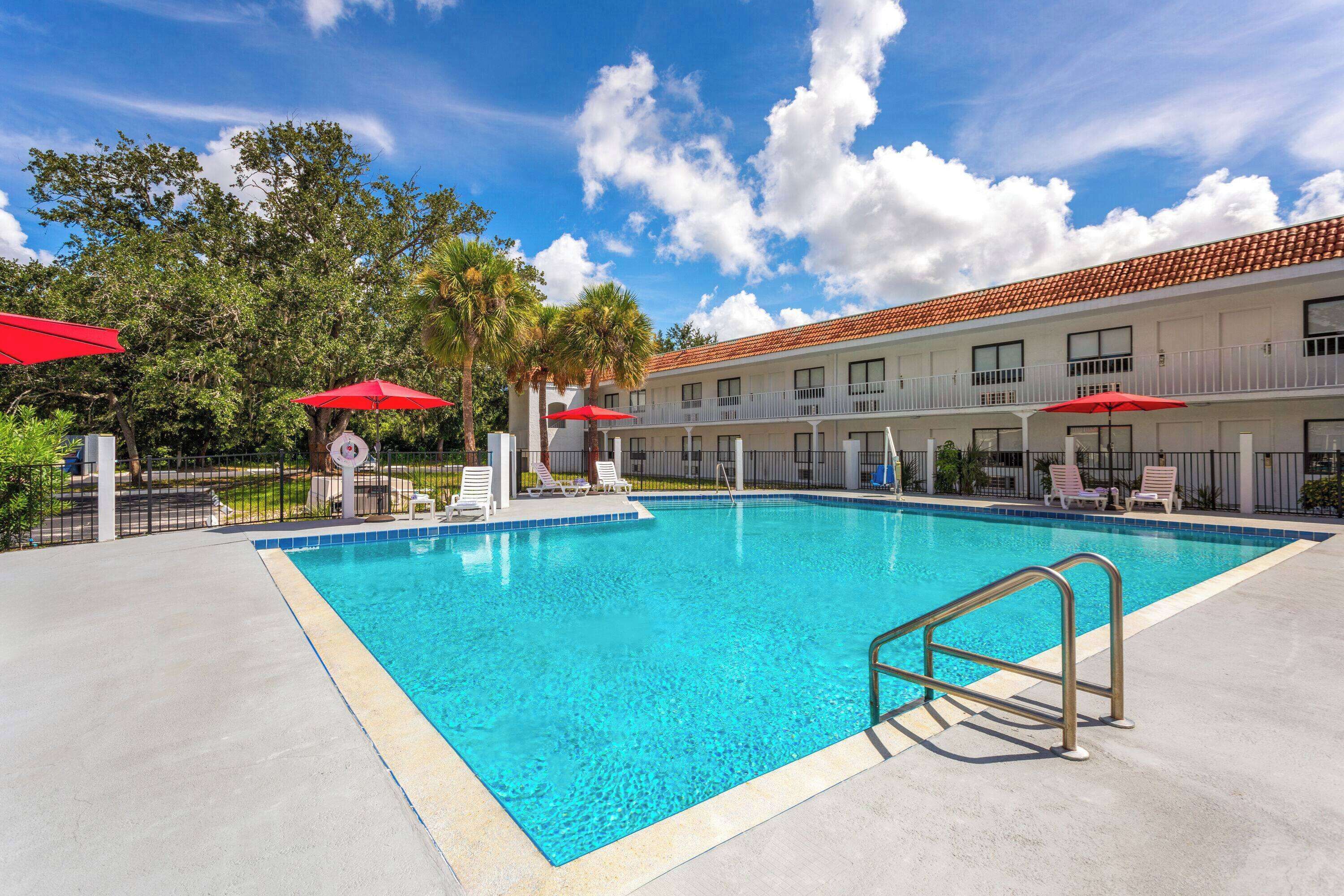 A swimming pool with a clear blue water, surrounded by lounge chairs and red umbrellas, with a building in the background.