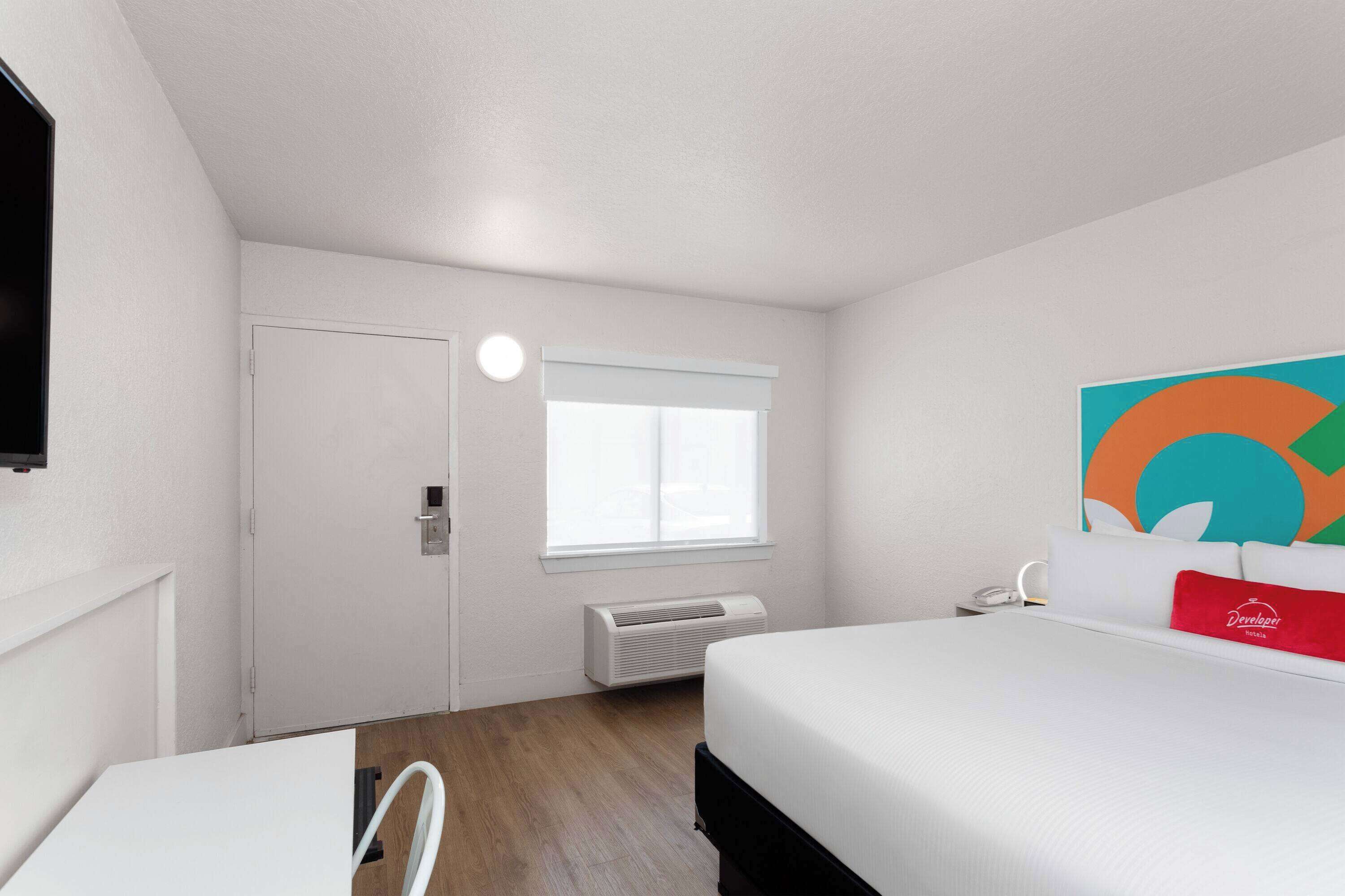 A hotel room with a bed, a desk, a chair, a window with blinds, an air conditioner, and a colorful artwork on the wall.