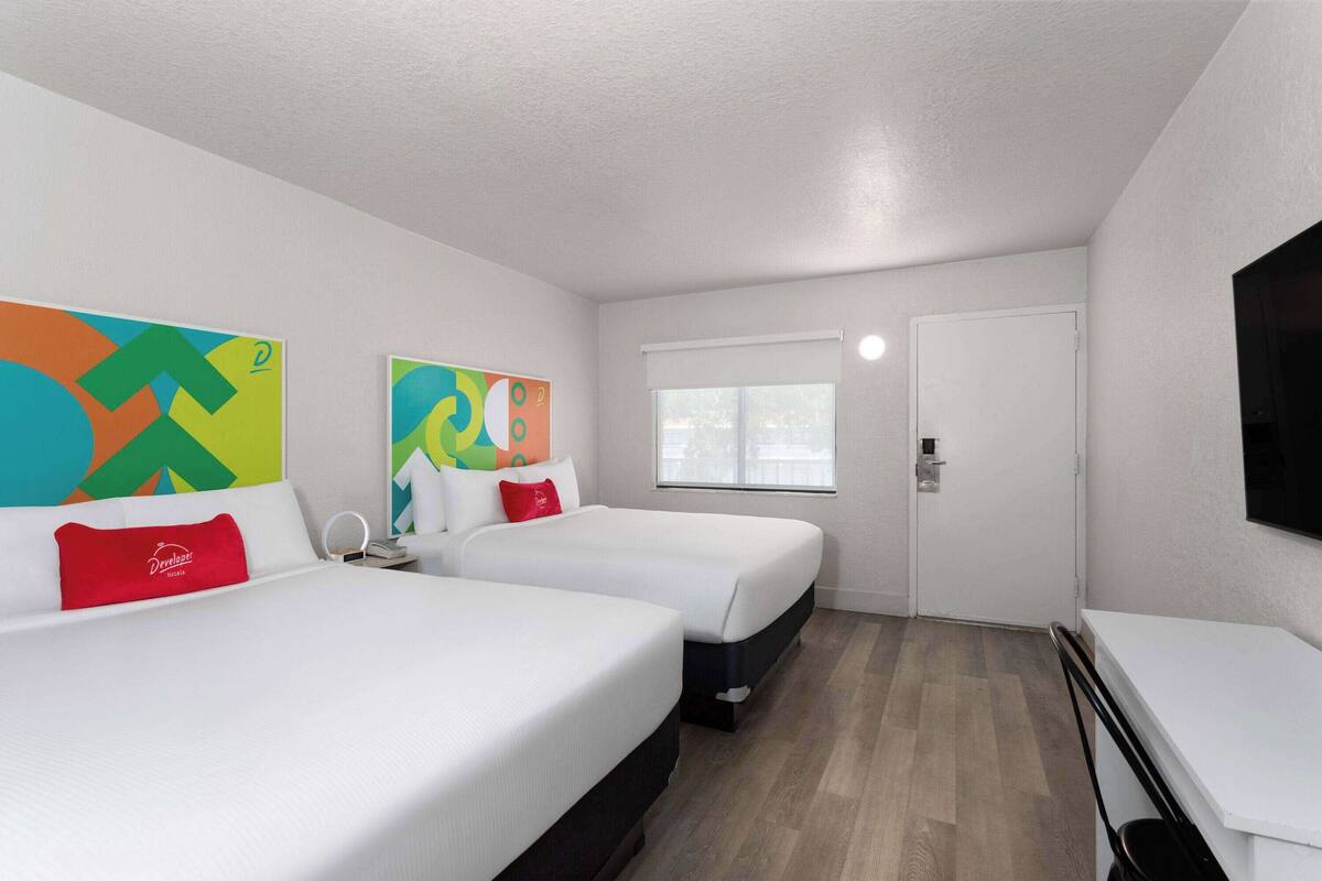 A hotel room with two beds, a desk, a chair, a TV, and artwork on the walls.