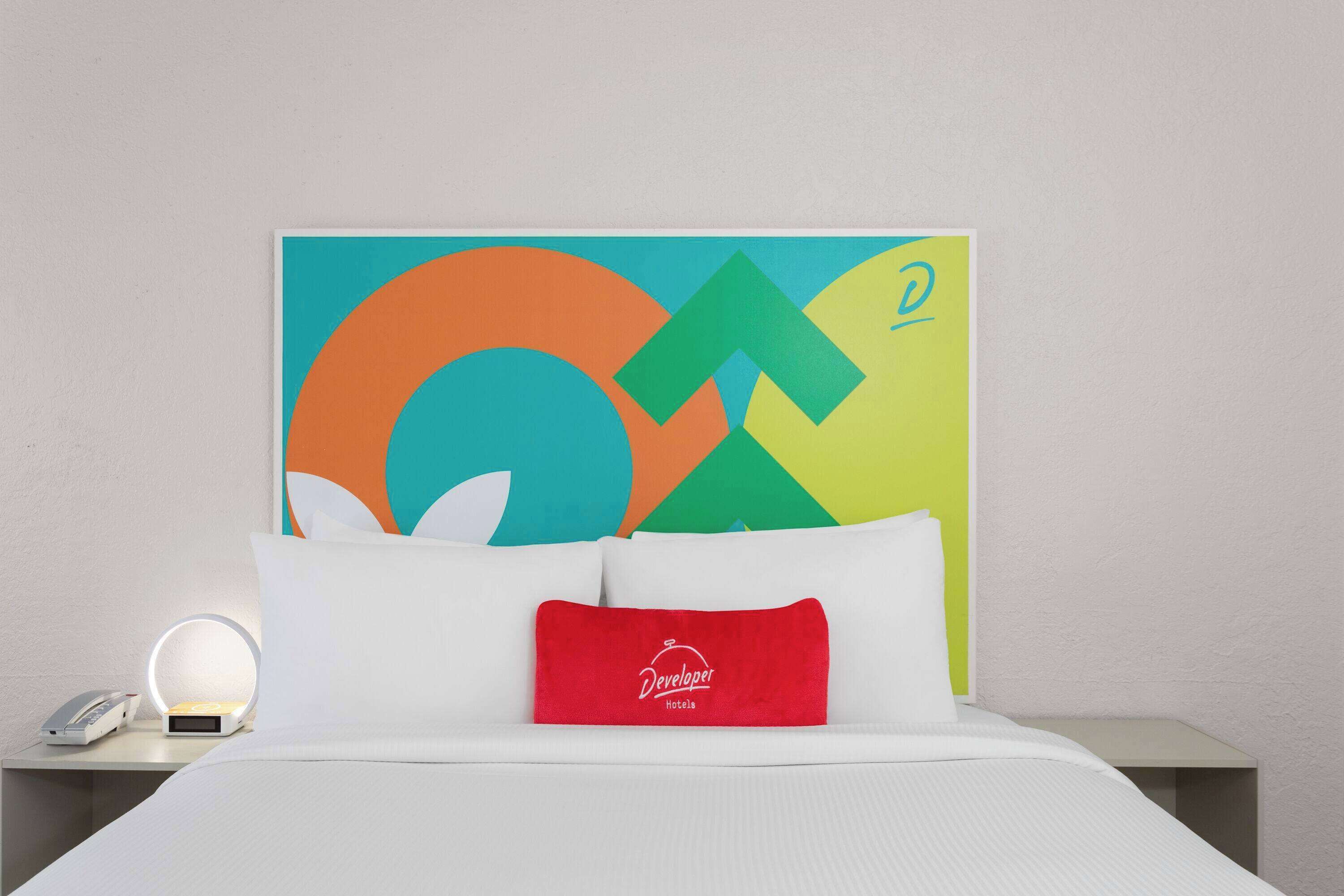 A hotel room with a bed, a nightstand with a lamp, and a colorful abstract painting on the wall.
