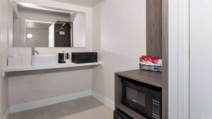 A modern bathroom with a white sink, a mirror, a towel rack, a microwave, and a shelf with toiletries. - Travelodge by Wyndham Orlando Lake Buena Vista South (Kissimmee)