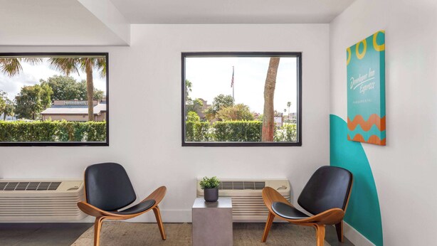 A modern waiting area with two black chairs, a small table with a plant, and a large window offering a view of trees and buildings. - Travelodge by Wyndham Orlando Lake Buena Vista South (Kissimmee)