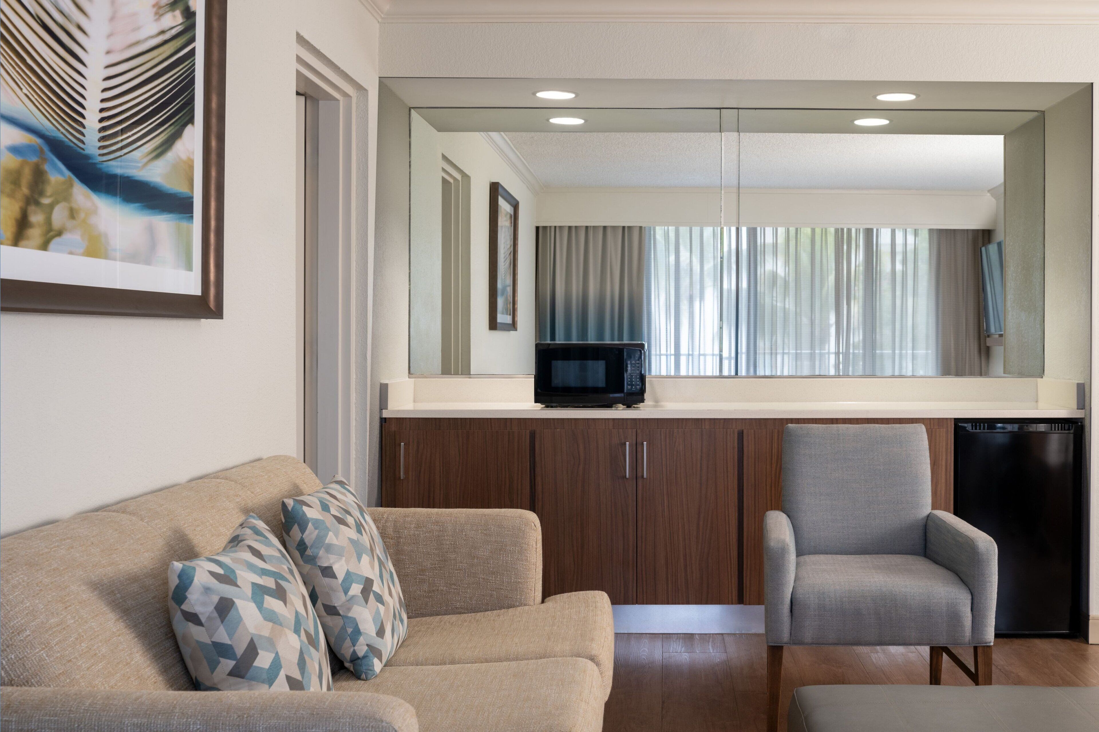 A modern living room with a beige sofa, two patterned pillows, a grey armchair, and a kitchen area with wooden cabinets, a microwave, and a dishwasher.
