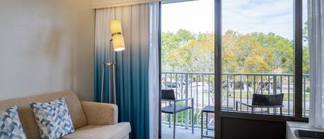 A hotel room with a sofa, two patterned pillows, a floor lamp, and a view of an outdoor balcony with chairs and trees.