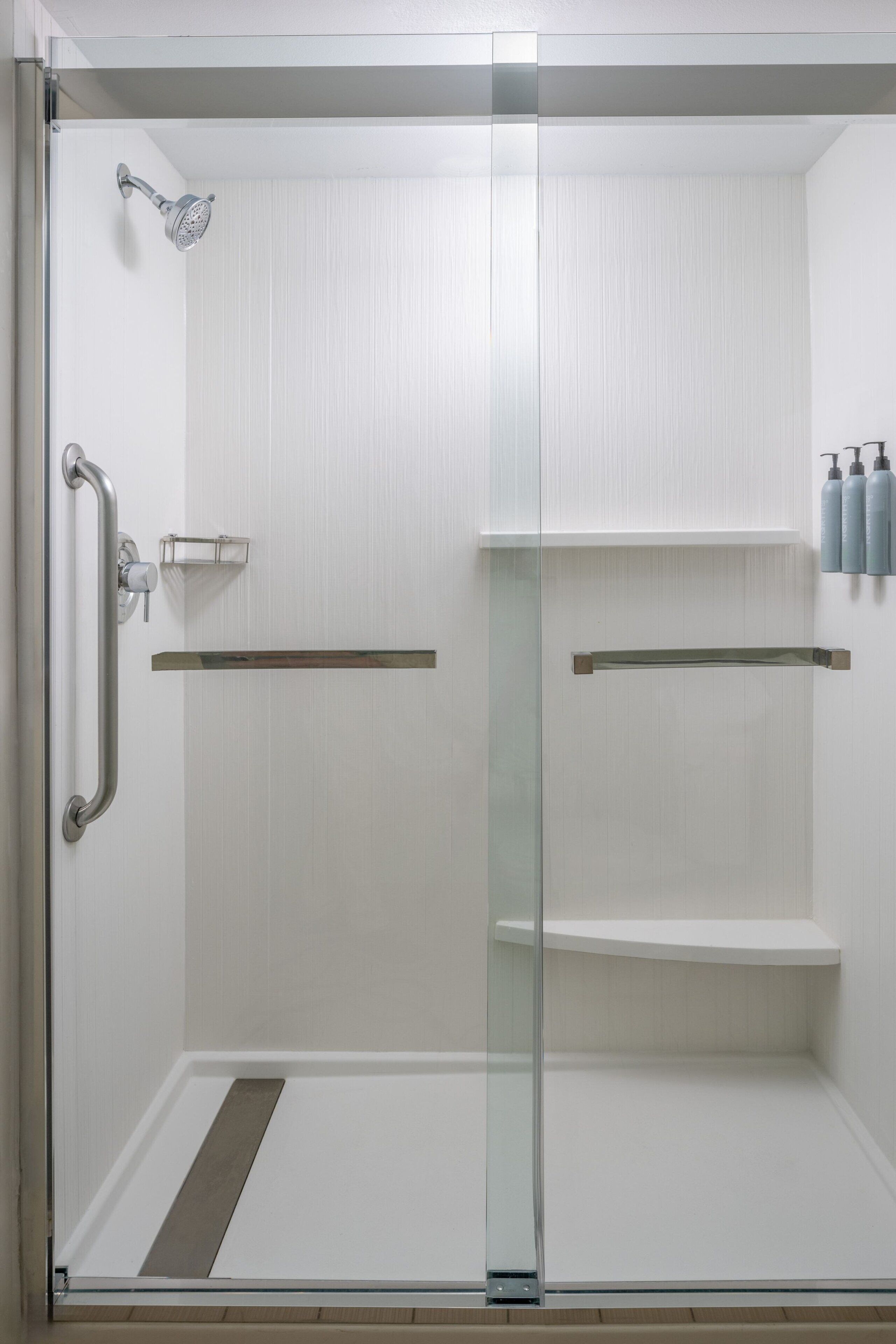 A glass-enclosed shower with a handheld showerhead, a grab bar, and a shelf with toiletries.