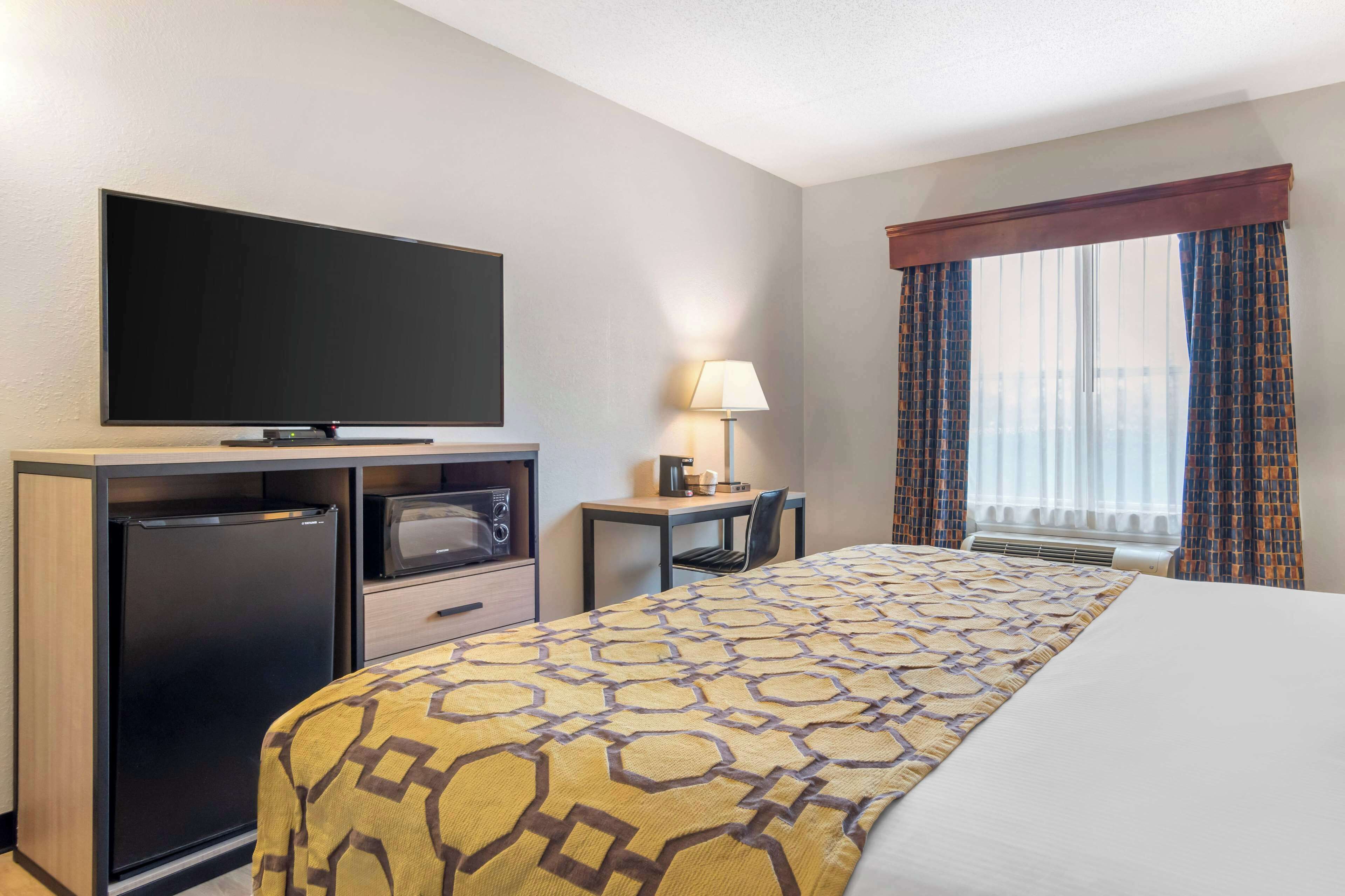 A hotel room with a bed, a flat-screen TV, a refrigerator, a microwave, a desk with a lamp, and a window with curtains.