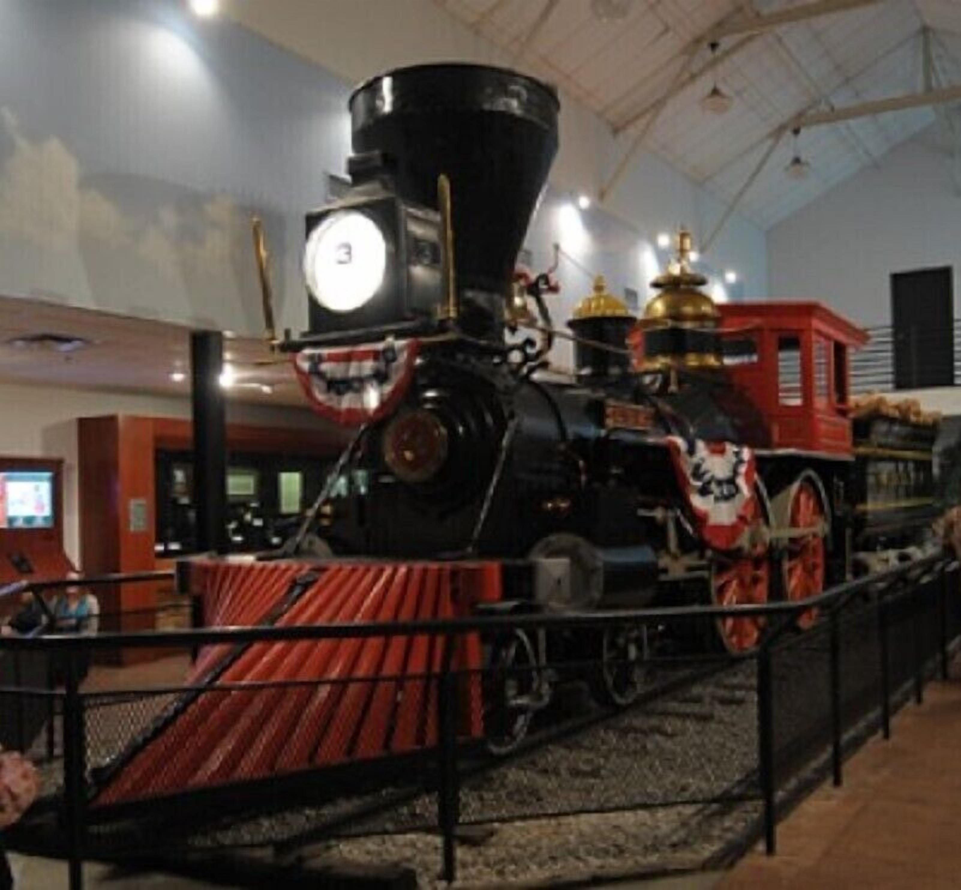 Una locomotora de vapor en exhibición, con un frente rojo, un reloj grande y una cúpula dorada en la parte superior.