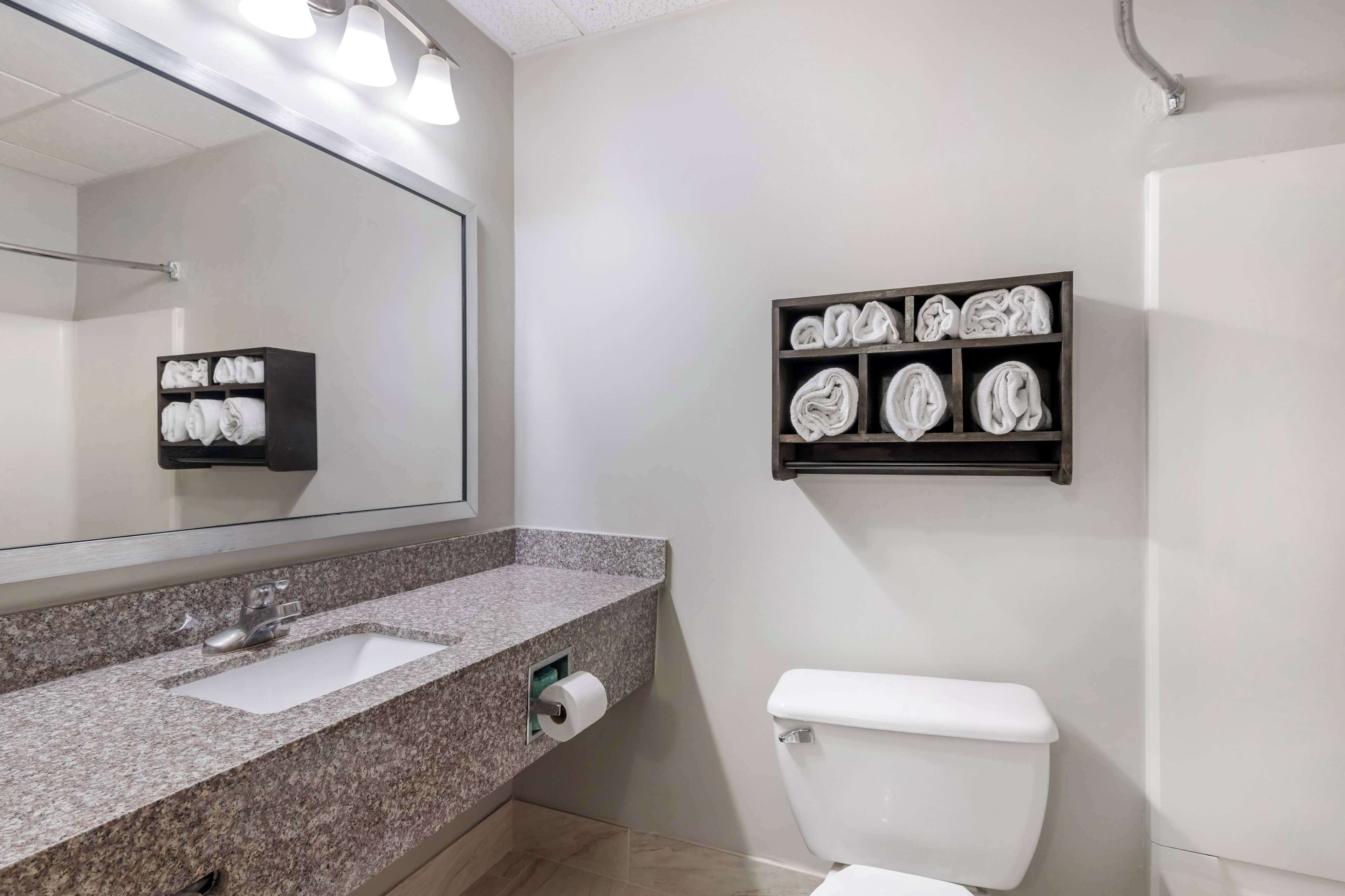 A bathroom with a granite countertop sink, a toilet, a towel rack with neatly folded towels, and a mirror with lights above it.