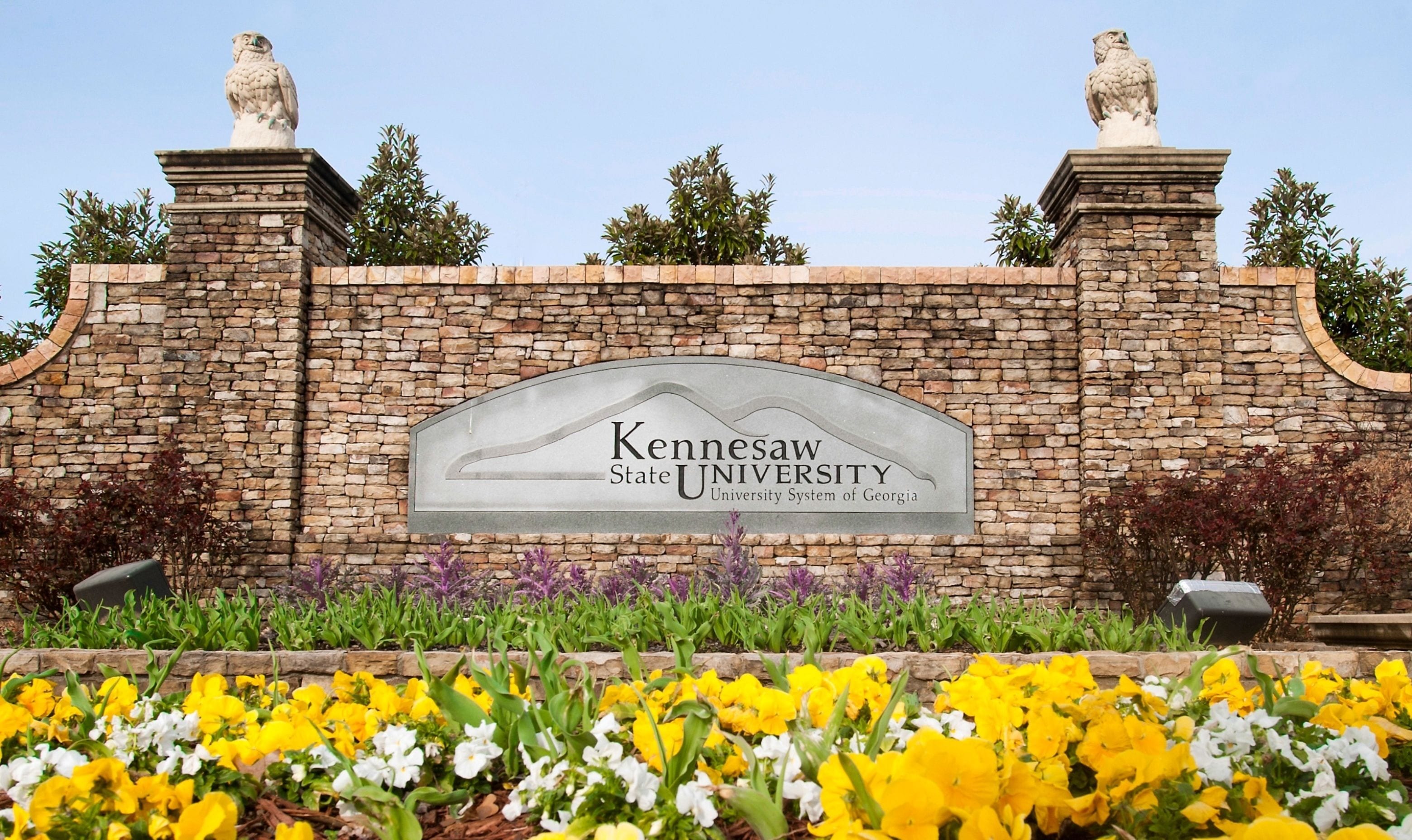 A stone wall with a sign for Kennesaw State University, surrounded by a garden with yellow and white flowers.