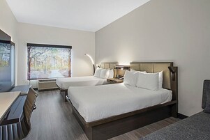 Habitación de hotel con dos camas, un escritorio y un ventanal con vistas a árboles.