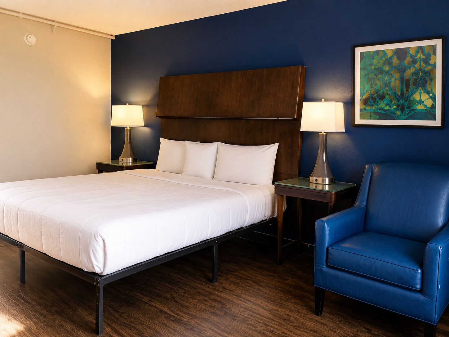 Ein Hotelzimmer mit einem Bett, zwei Nachttischen mit Lampen, einem blauen Sessel und einem Bild an der Wand.
