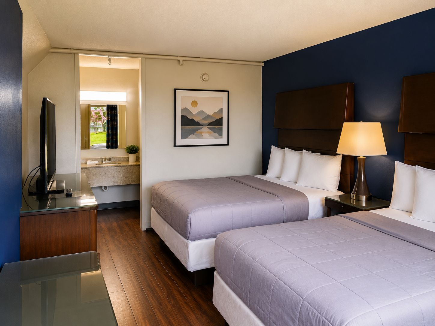 A hotel room with two beds, a television, a bathroom, and a painting on the wall.