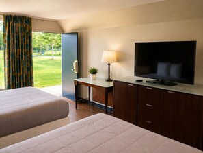 A hotel room with a bed, a desk with a television, and a view of a green outdoor area.