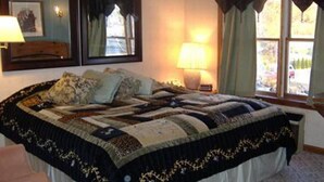 A bedroom with a large bed, a bedside lamp, a mirror, and a window with curtains. - Sturbridge Country Inn (Fiskdale)