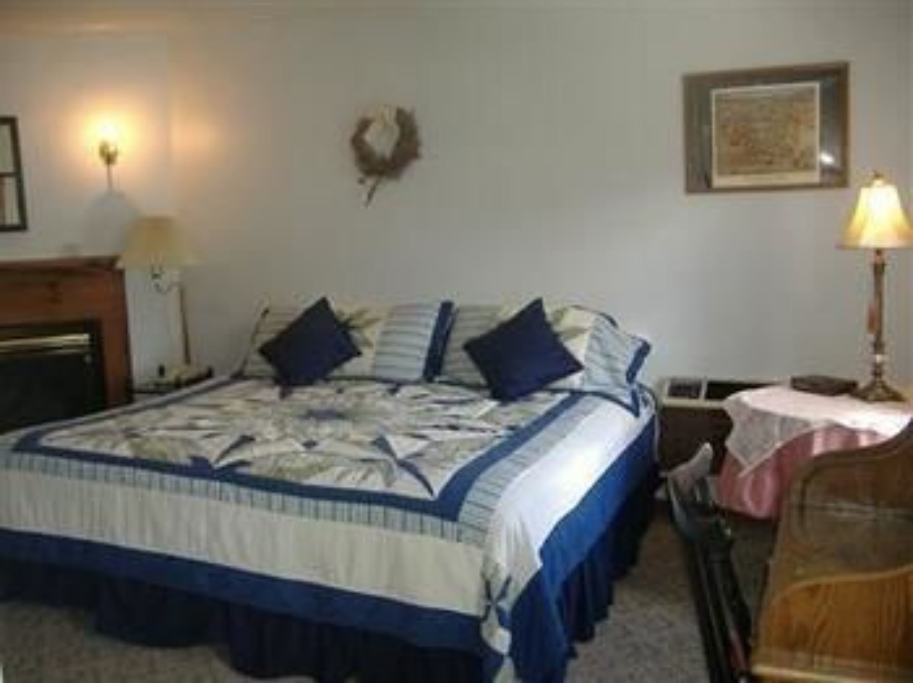 A bedroom with a bed, a nightstand, a lamp, a framed picture on the wall, and a small table with a tablecloth.
