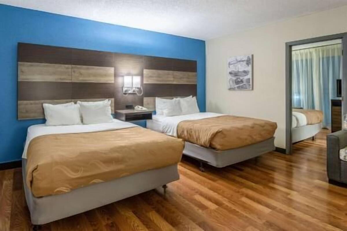 A hotel room with two beds, wooden flooring, a blue accent wall, a nightstand with a phone, and a painting.