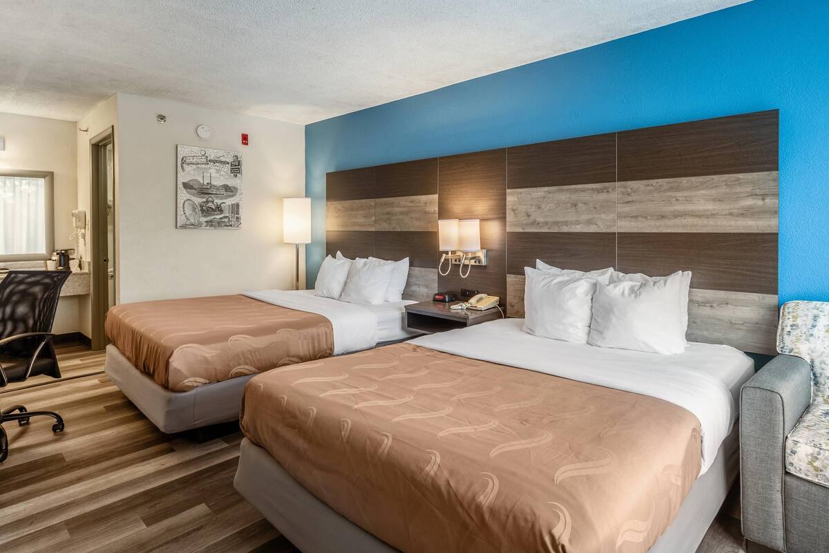 A hotel room with two beds, a desk, a chair, and a wall with a clock and artwork.