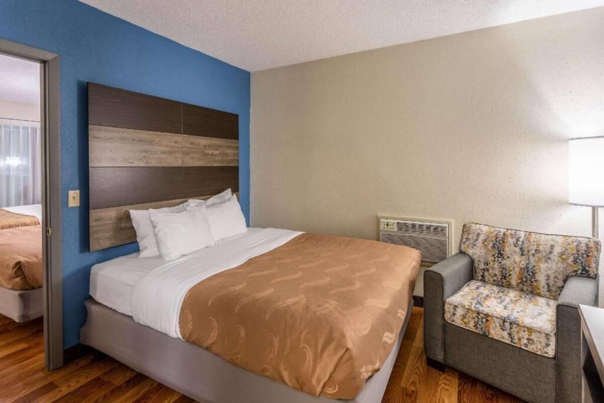 A hotel room with a bed, a chair, a heater, and a door leading to another room.