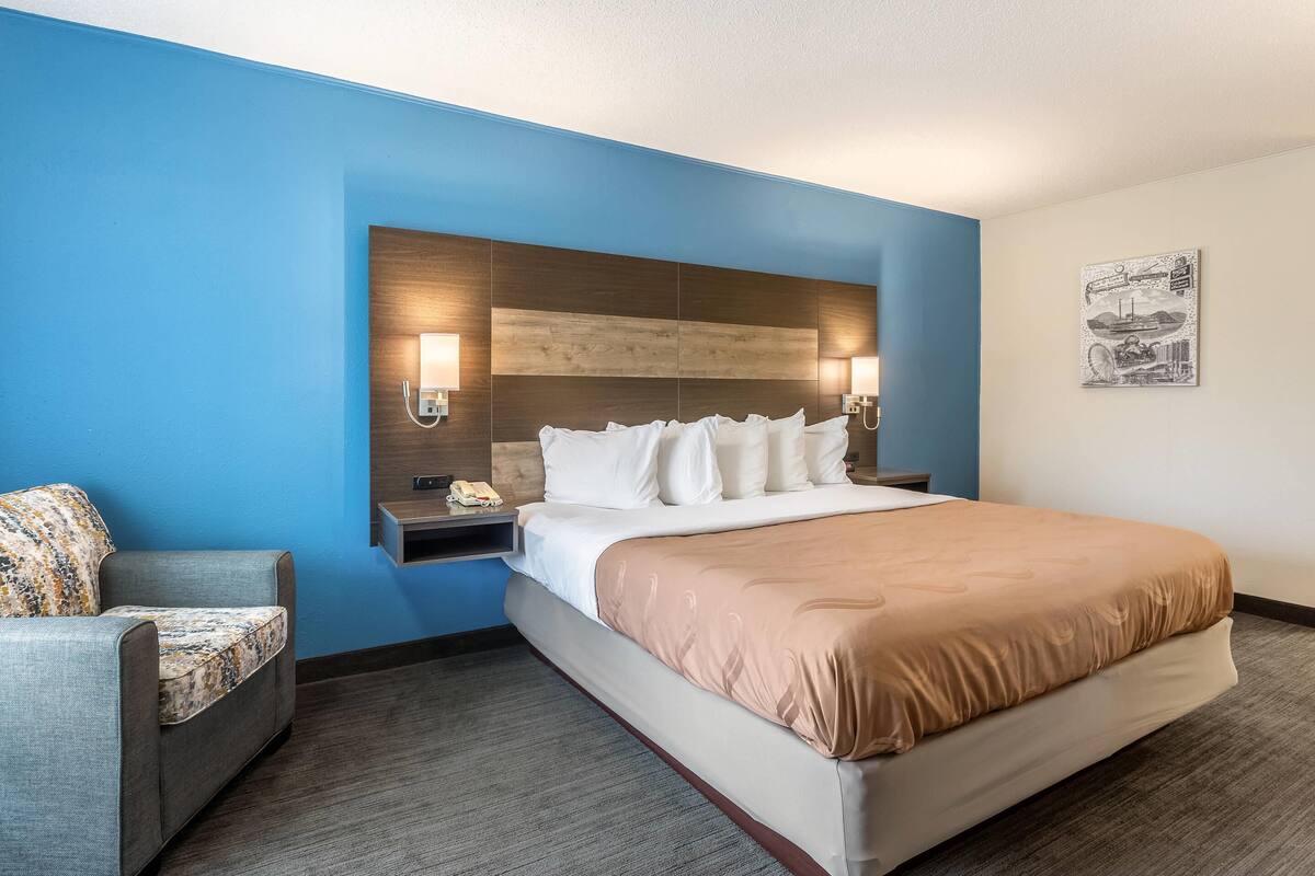 A hotel room with a bed, a nightstand, a chair, a wall-mounted light, and a picture on the wall.