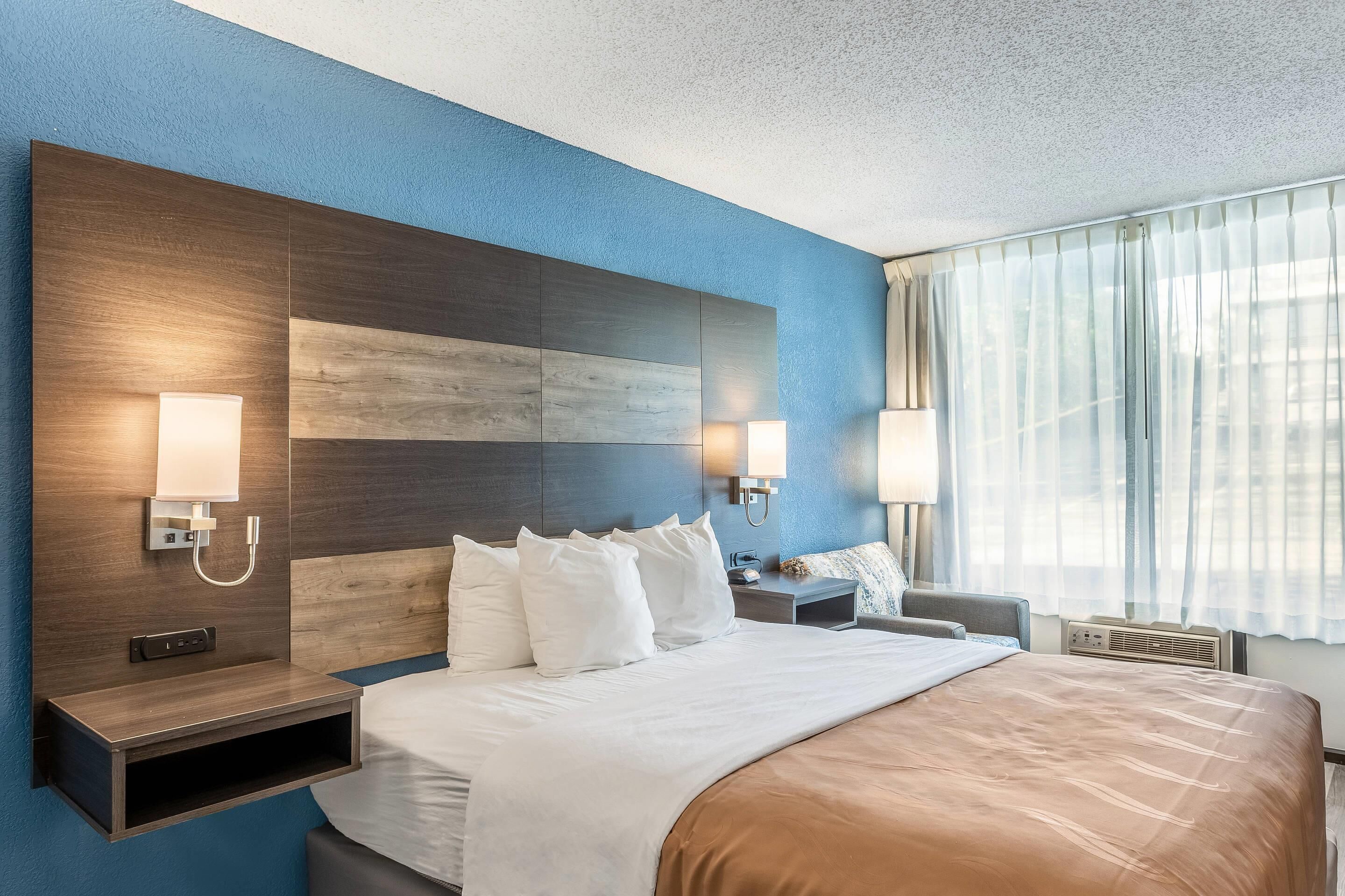 Ein Hotelzimmer mit einem großen Bett, Nachttischlampen, einem Beistelltisch und einem Fenster mit durchscheinenden Vorhängen.