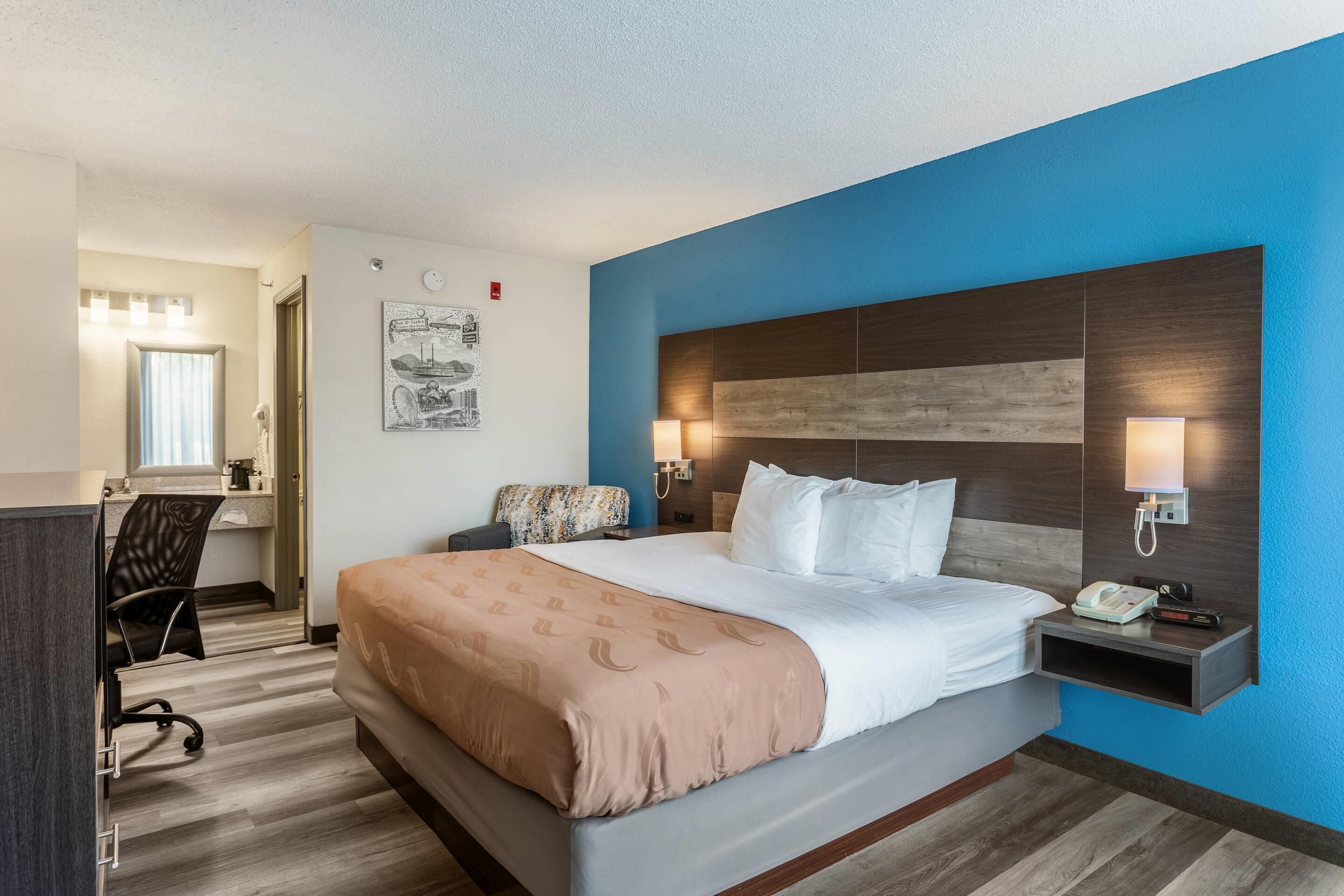 Ein Hotelzimmer mit Bett, Nachttisch, Lampe, Schreibtisch, Stuhl und einem an der Wand befestigten Telefon.