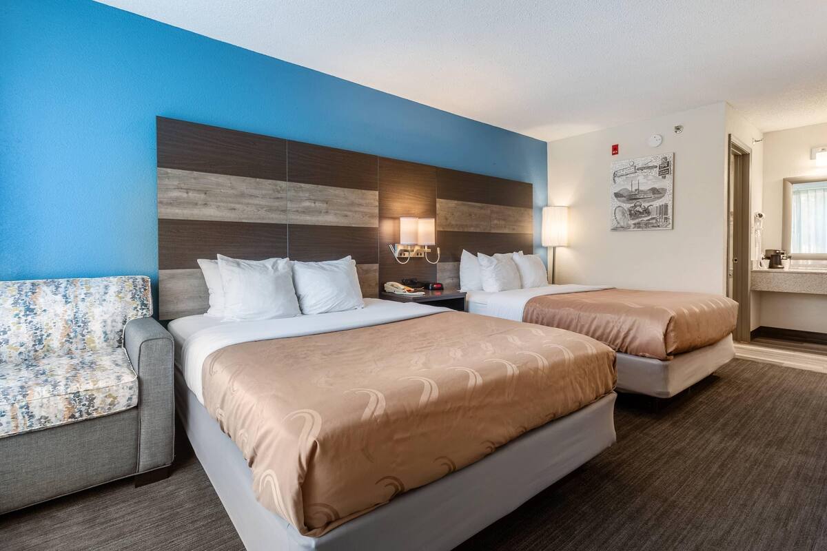 A hotel room with two beds, a sofa, a nightstand with a lamp, and a wall-mounted artwork.