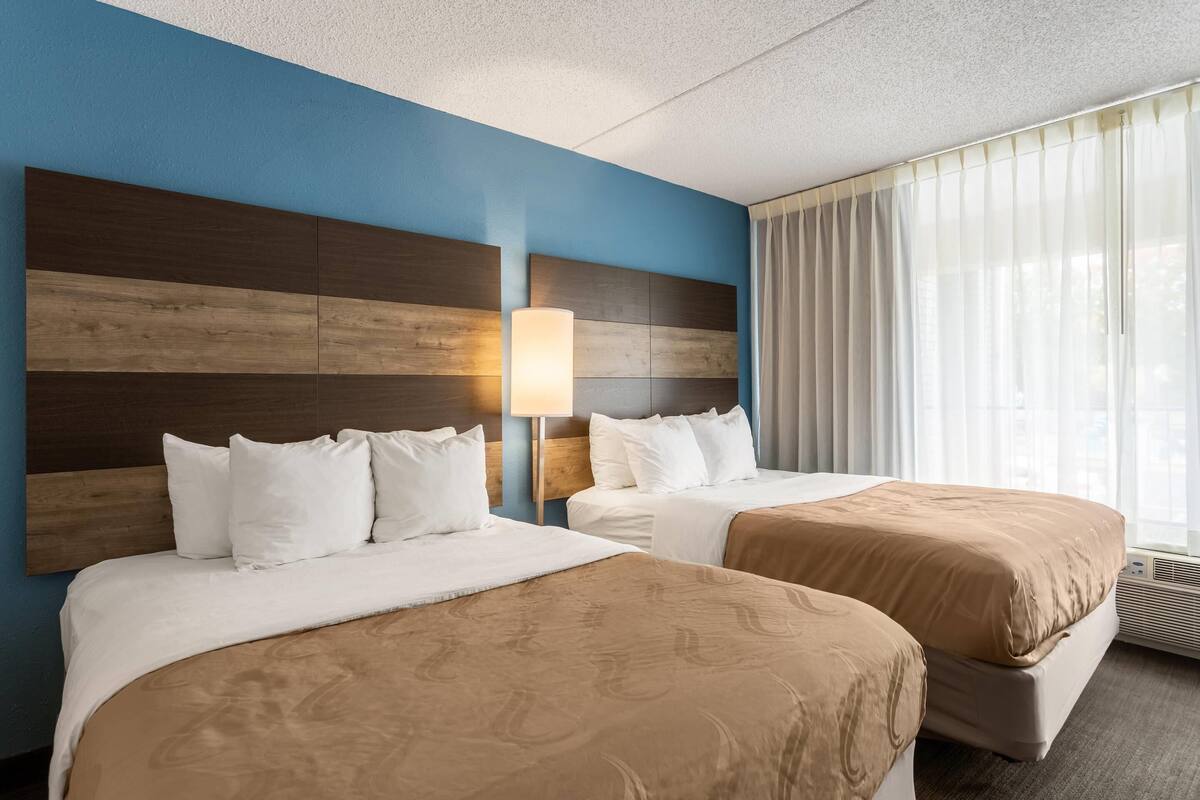 A hotel room with two beds, a large window with curtains, a wall-mounted lamp, and a ceiling with a textured finish.