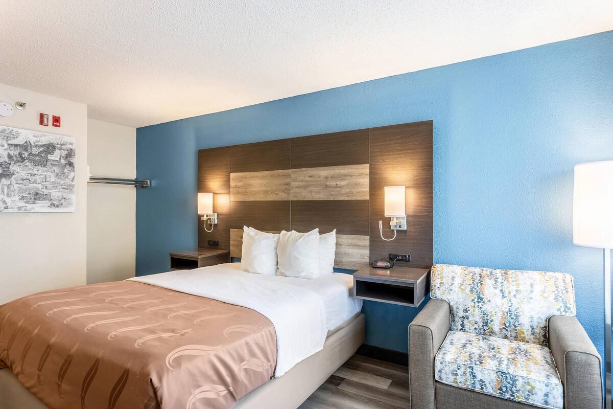 A hotel room with a bed, two bedside tables, a chair, a headboard with wall art, and a wall-mounted lamp.