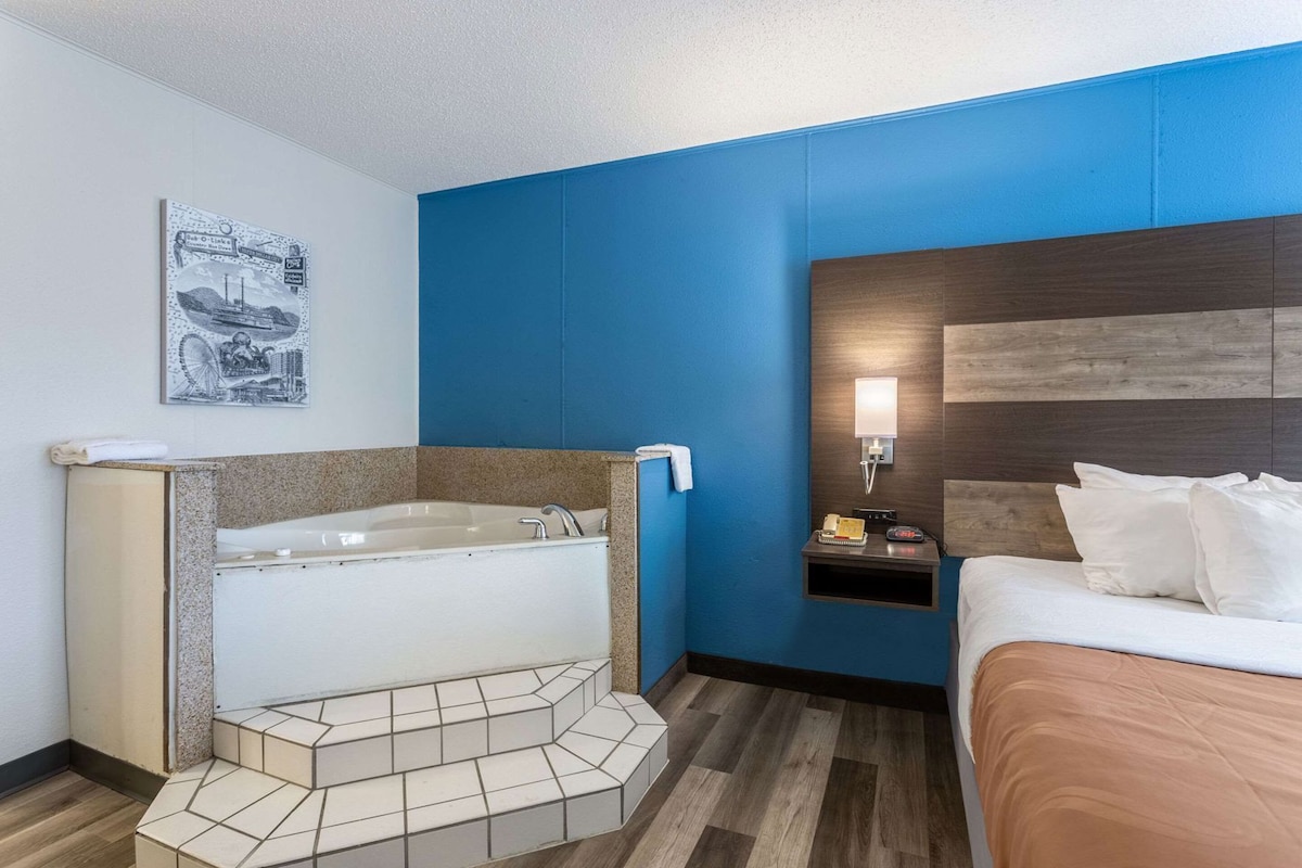 A hotel room with a bathtub, a bed, a nightstand, and a wall-mounted light.