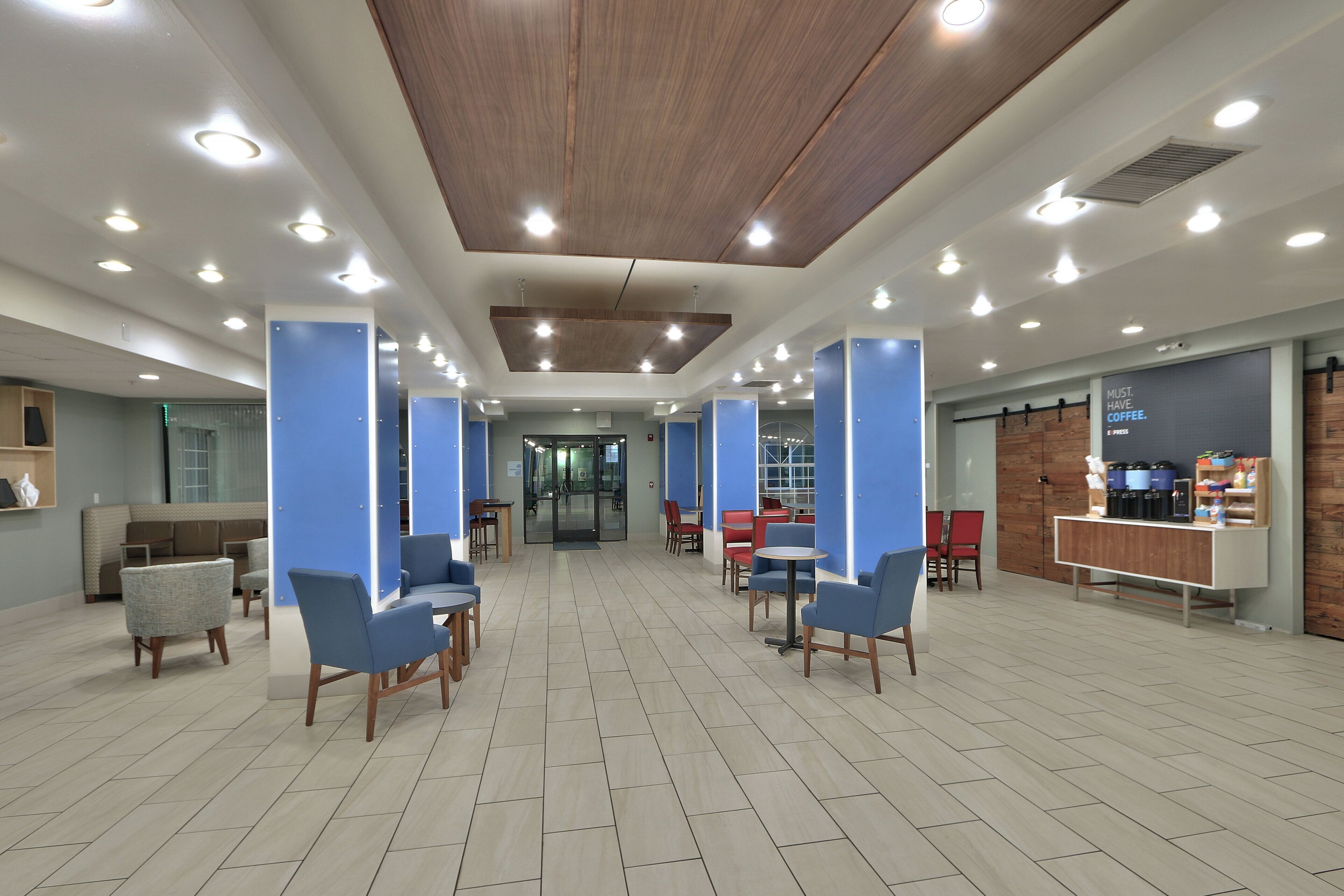 A spacious lobby with a modern design, featuring a central seating area with chairs and tables, a reception desk, and a coffee station.
