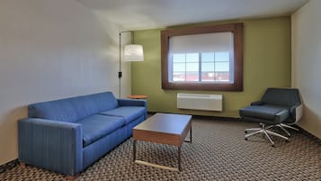Habitación de hotel con un sofá azul, una mesa pequeña, una silla, una ventana con persianas y una lámpara.