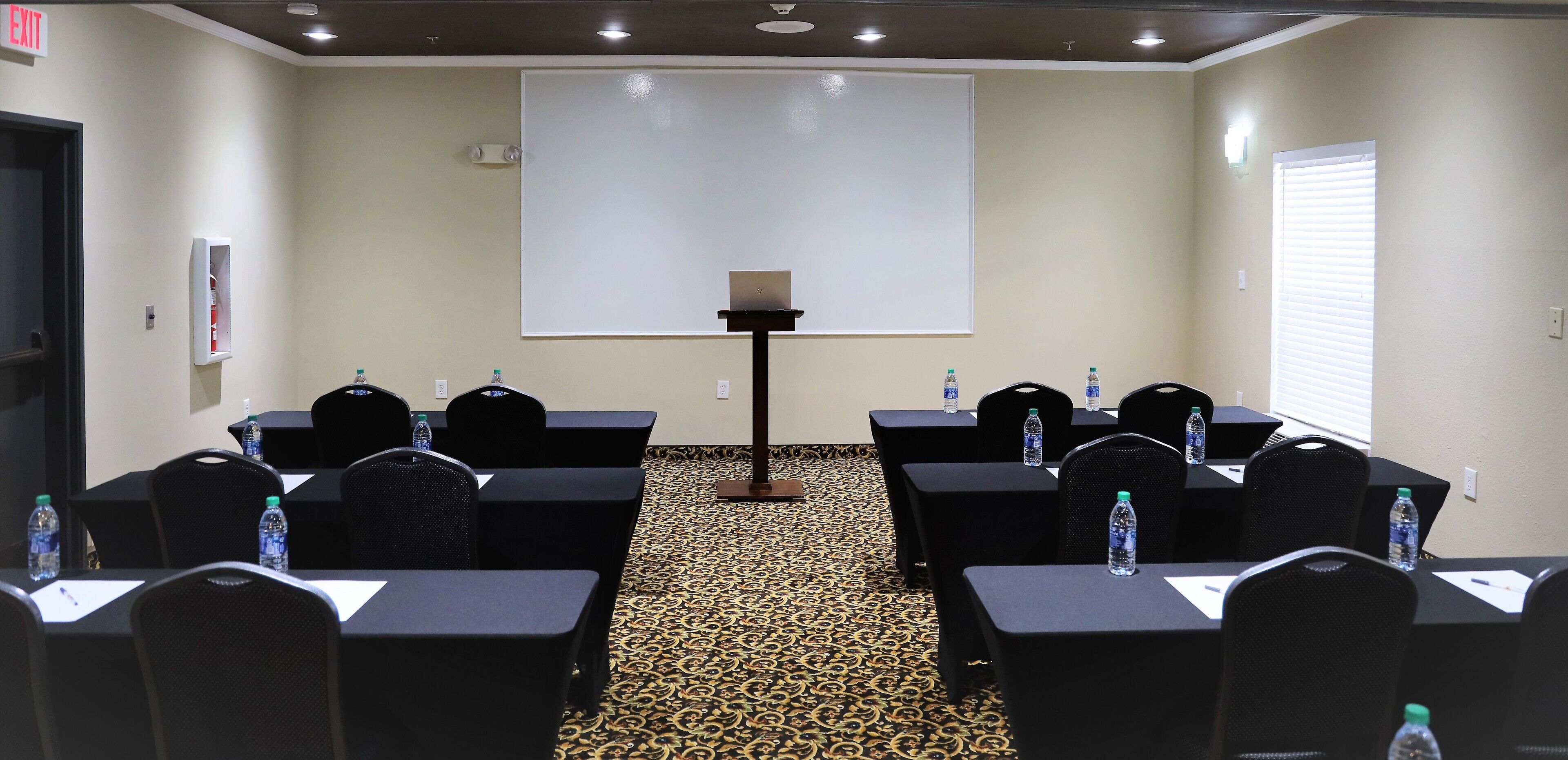 Una sala de conferències amb pantalla de projector, taules, cadires, ampolles d'aigua i un senyal d'emergència.