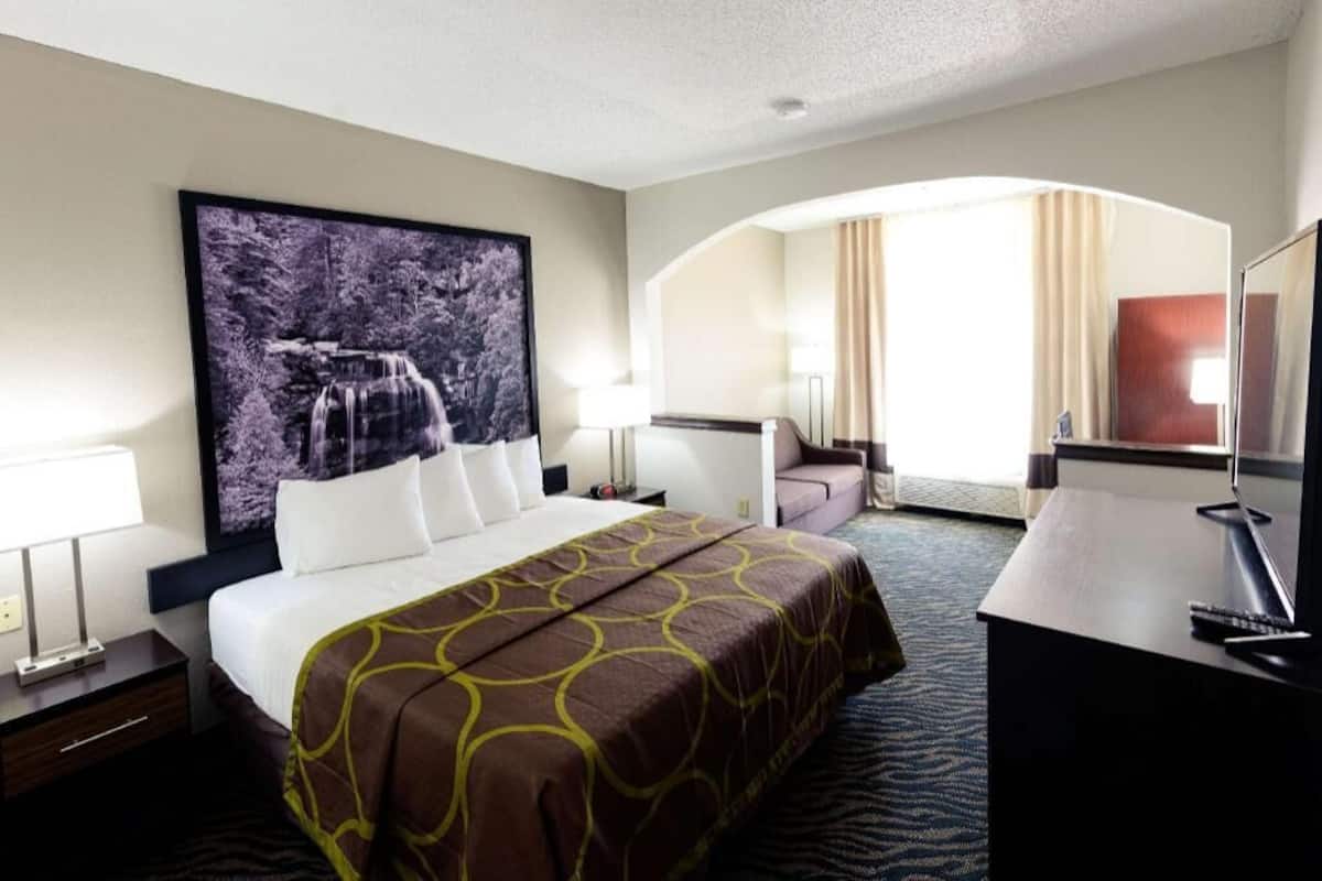 Ein Hotelzimmer mit Bett, Schreibtisch, Fernseher, einem großen Fenster mit Vorhängen und einem Bild von einem Wasserfall.