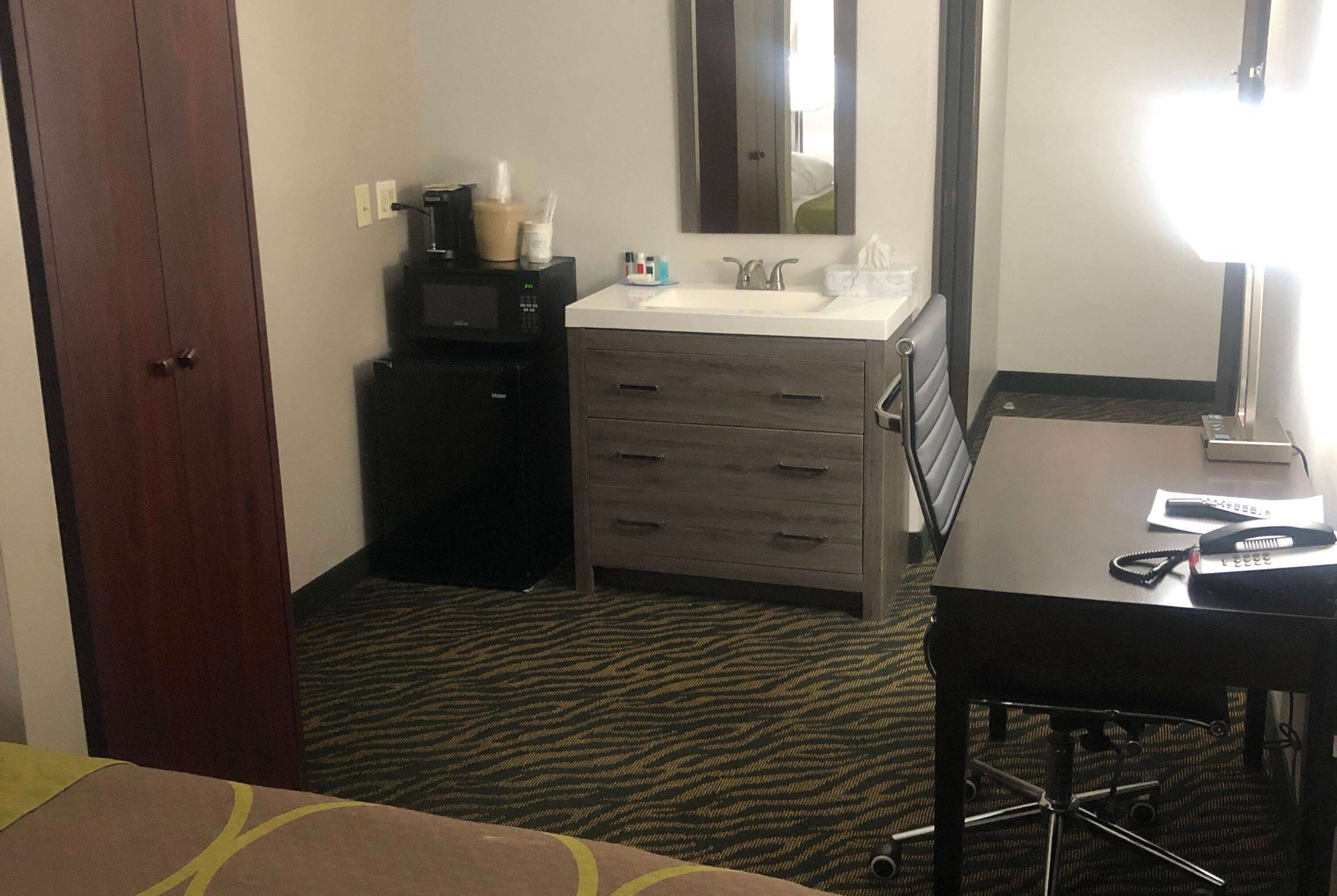 Una habitación de hotel con cama, escritorio con lámpara, cómoda, espejo y microondas.