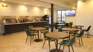 Un café avec des tables rondes et des chaises vertes, un coin cuisine équipé d’appareils électroménagers et un écran de télévision fixé au mur.
