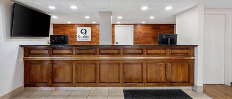 A reception desk with a 'Quality by Choice Hotels' sign, wooden paneling, a flat-screen TV, and a black carpet.