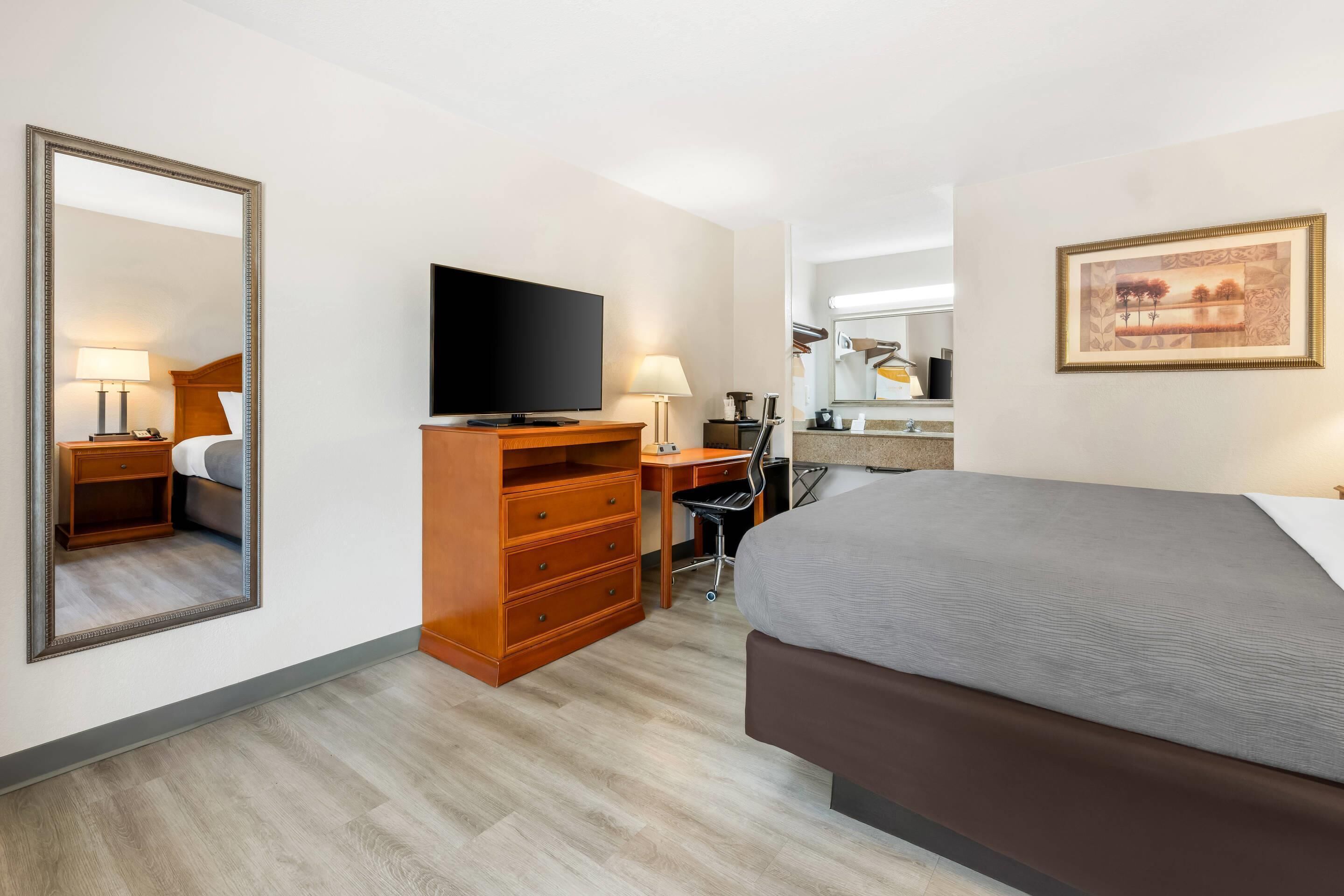 A hotel room with a bed, a desk with a computer, a television, a mirror, and a framed picture on the wall.