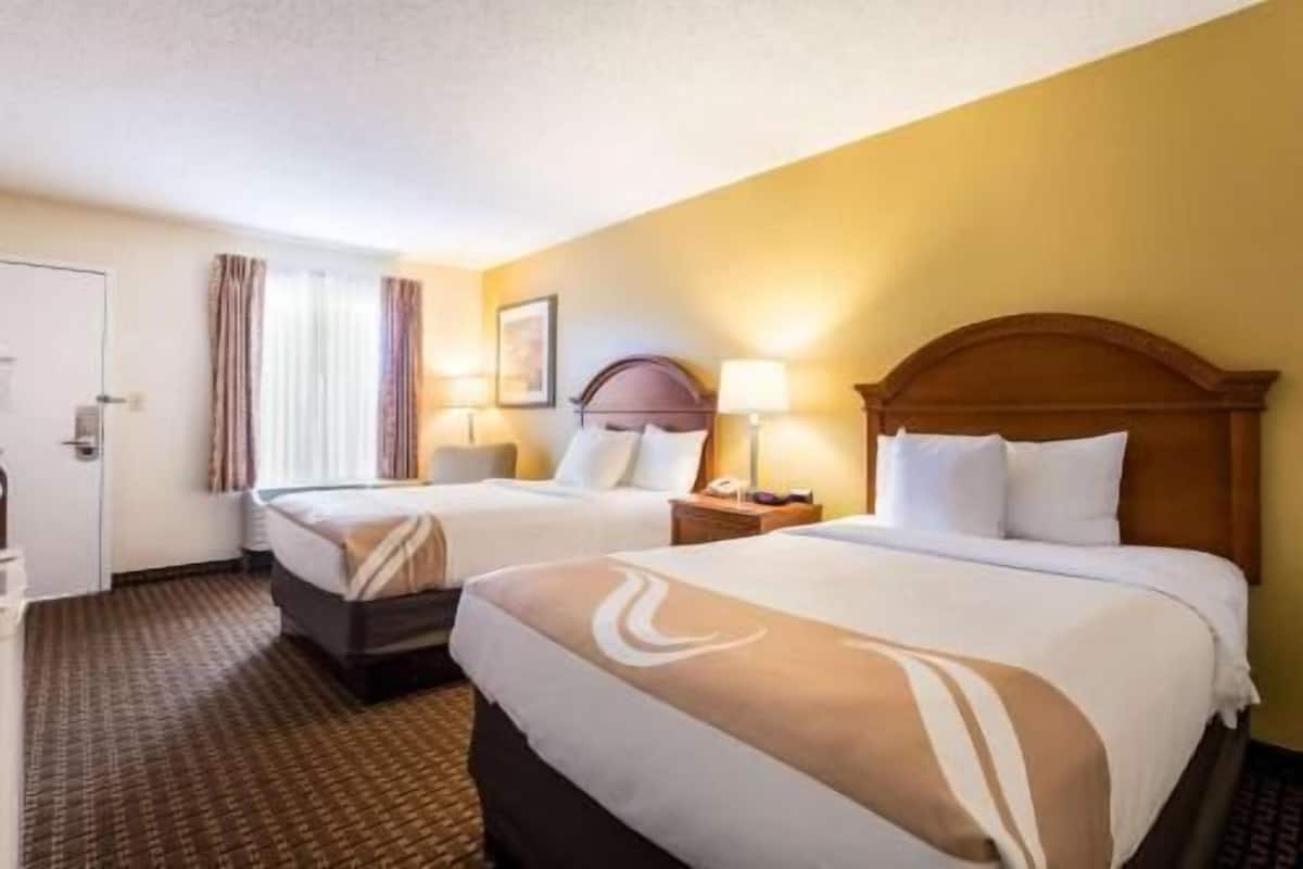 A hotel room with two beds, a headboard, bedside tables, a lamp, a window with curtains, and a door.