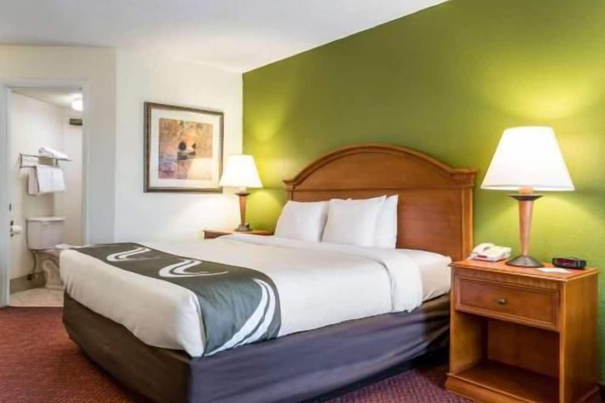 A hotel room with a bed, a nightstand with a lamp, a framed picture on the wall, and a bathroom visible in the background.