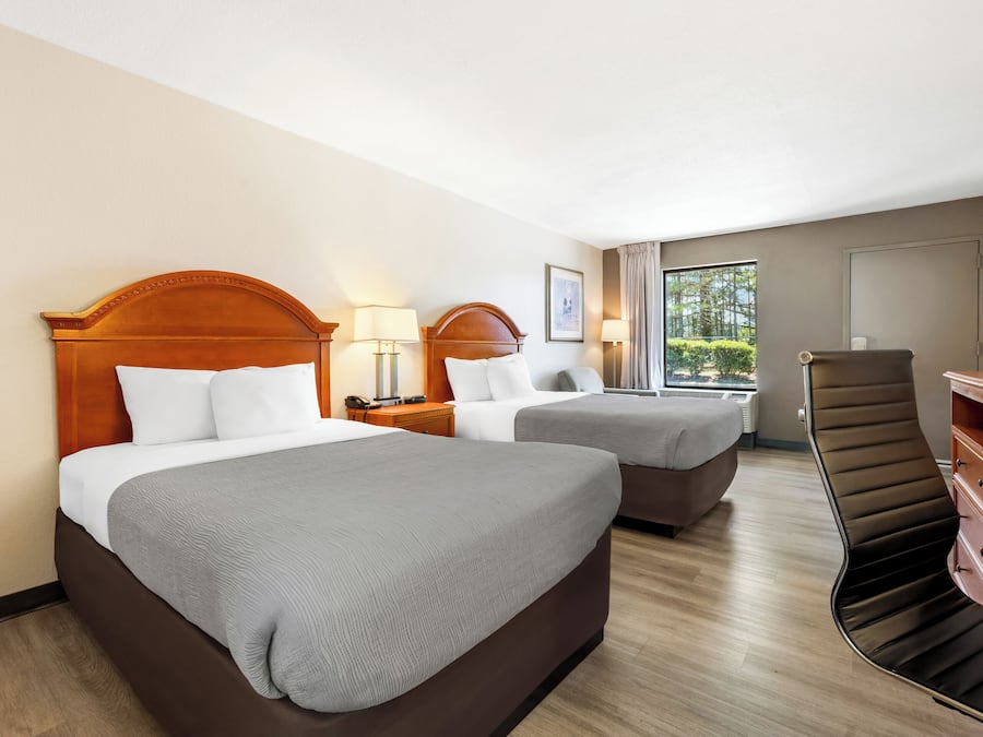 A hotel room with two beds, a desk, a chair, and a view of greenery outside.
