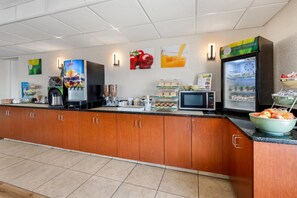 A self-service breakfast area with a coffee machine, juice dispenser, microwave, and refrigerated display.
