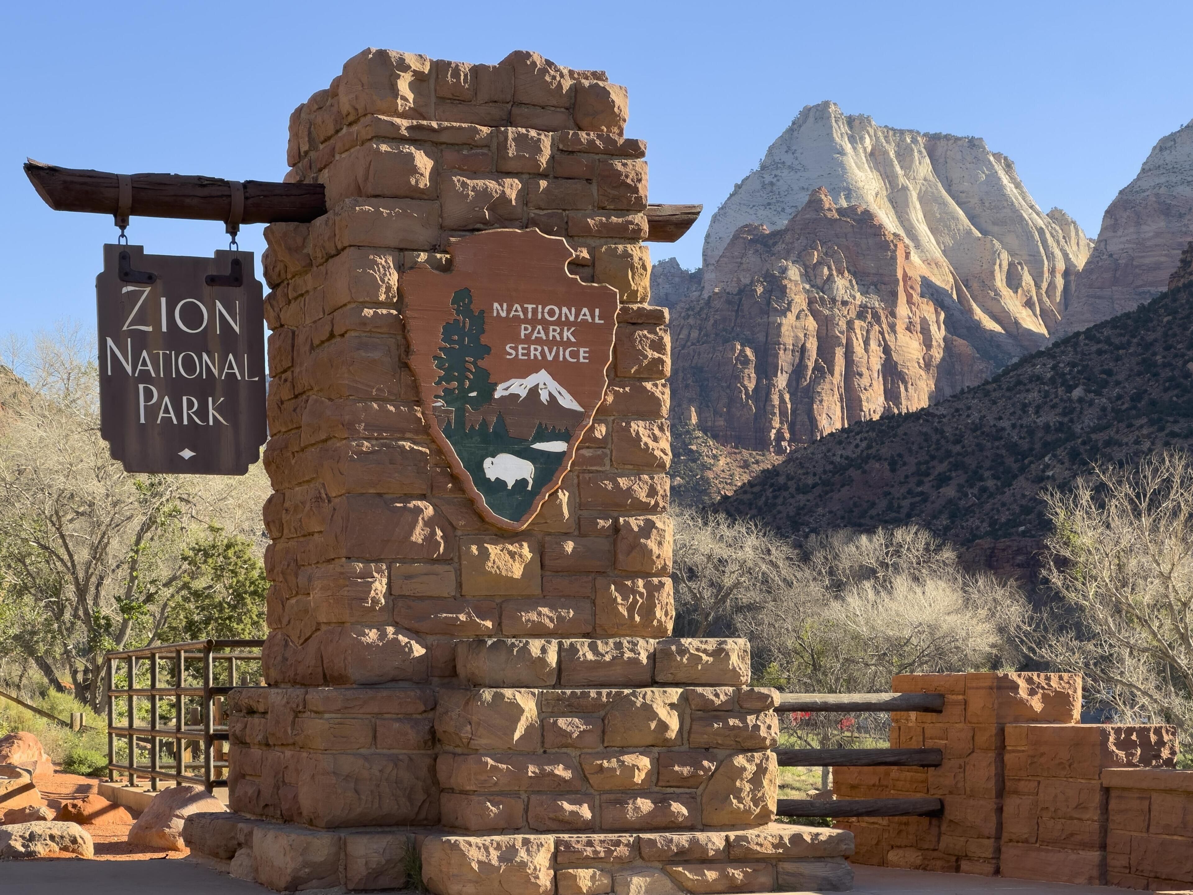 A stone sign with 'Zion National Park' and 'National Park Service' logos, a wooden gate, and a mountainous backdrop.