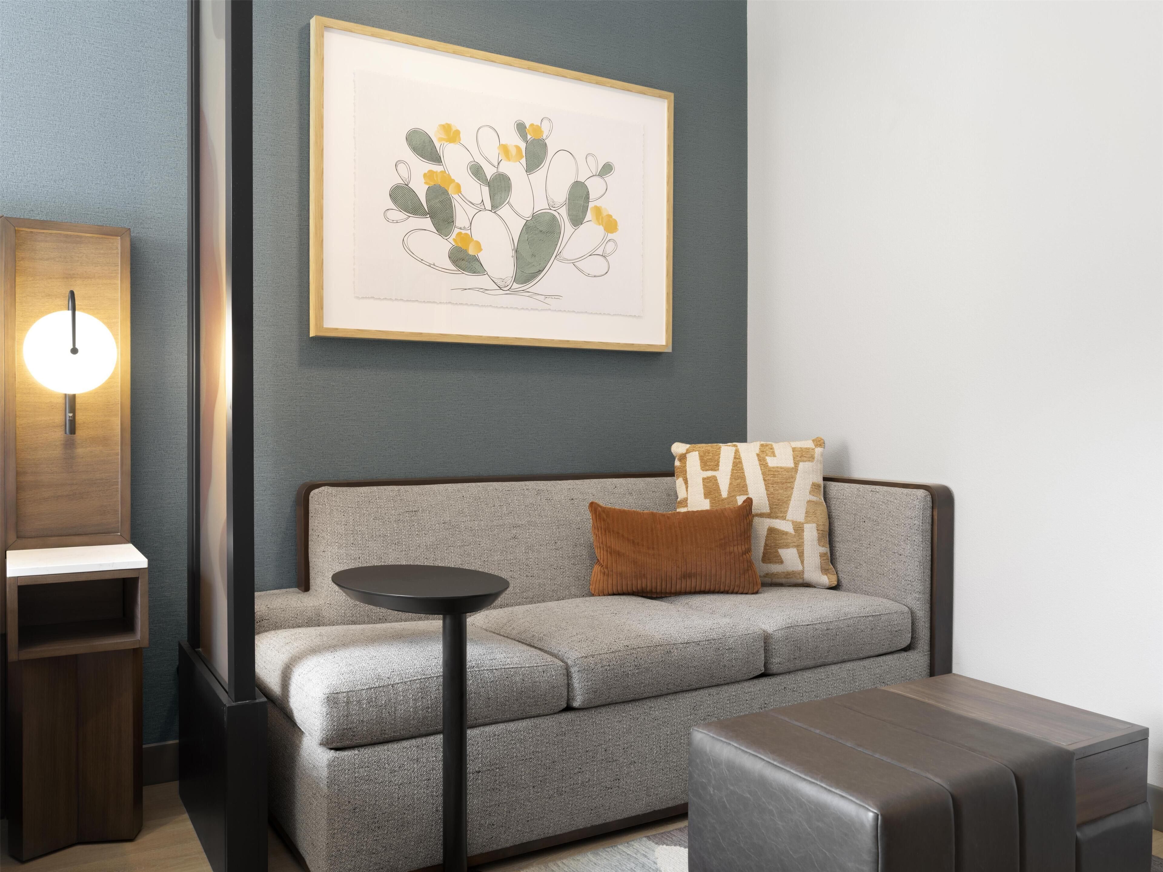 A modern living room with a grey sofa, a round side table, a framed artwork, and a wall-mounted light fixture.