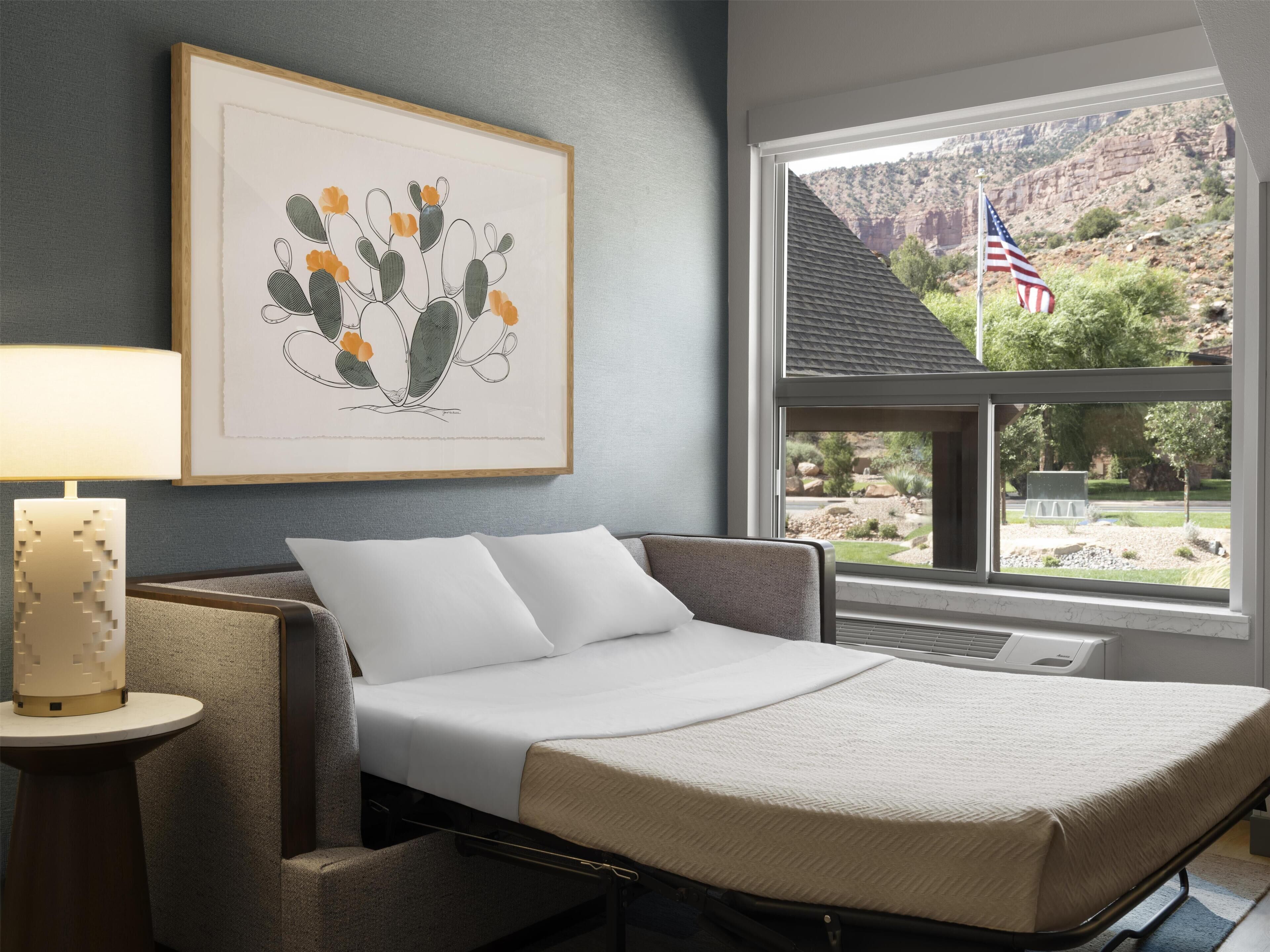 A bedroom with a bed, a framed cactus artwork, a lamp, and a window with a view of a mountainous landscape.