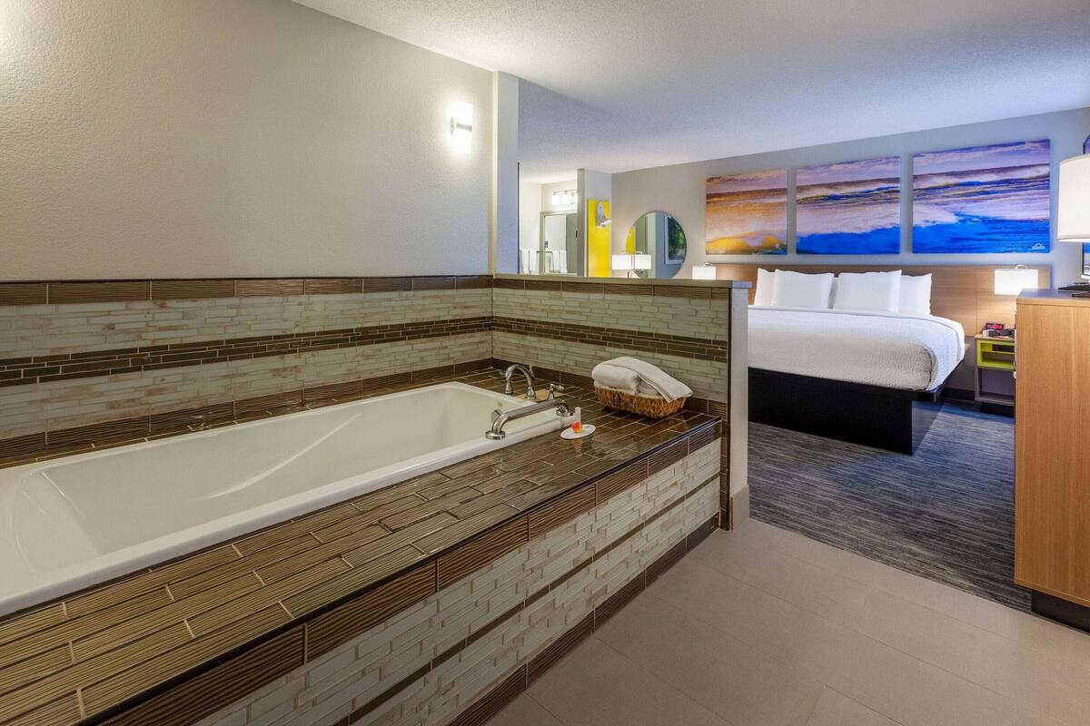 Une salle de bain d’hôtel moderne dotée d’une grande baignoire, d’une douche séparée et d’un lit offrant une vue sur le paysage urbain.